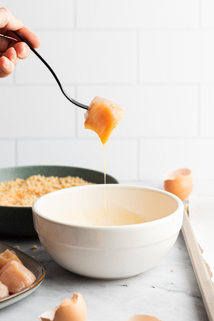 Fork holding chicken breast cube dipped in egg, letting excess drip off