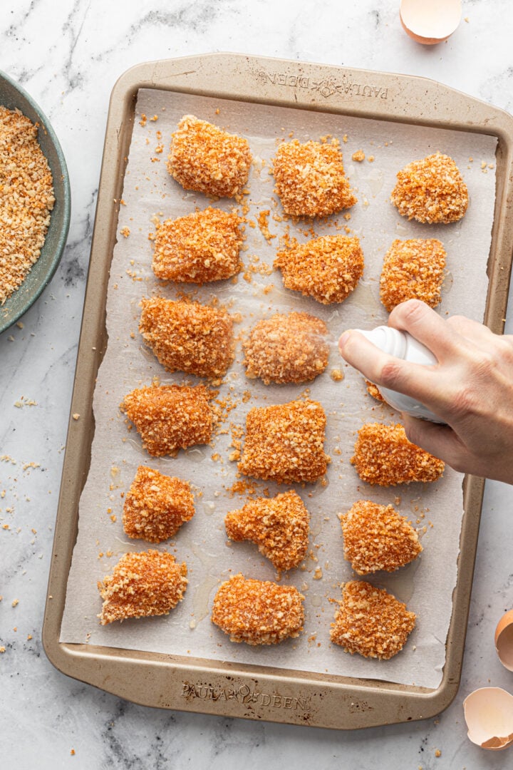 Cooking spray being sprayed onto baked panko nuggets on a lined sheet pan