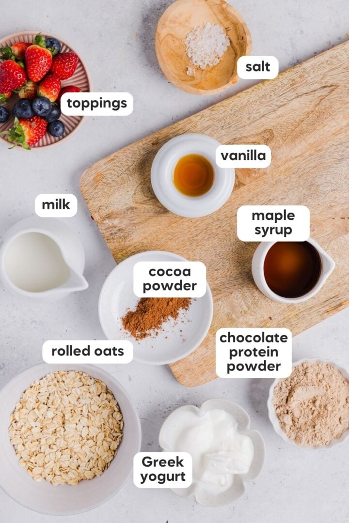 Overhead and labeled photo of ingredients in chocolate protein overnight oats, displayed in small bowls on a marble counter