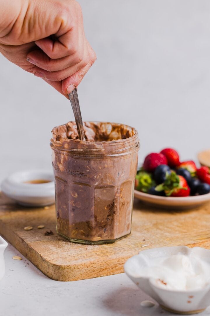 Women's hand stirring chocolate protein overnight oats in small glass jar