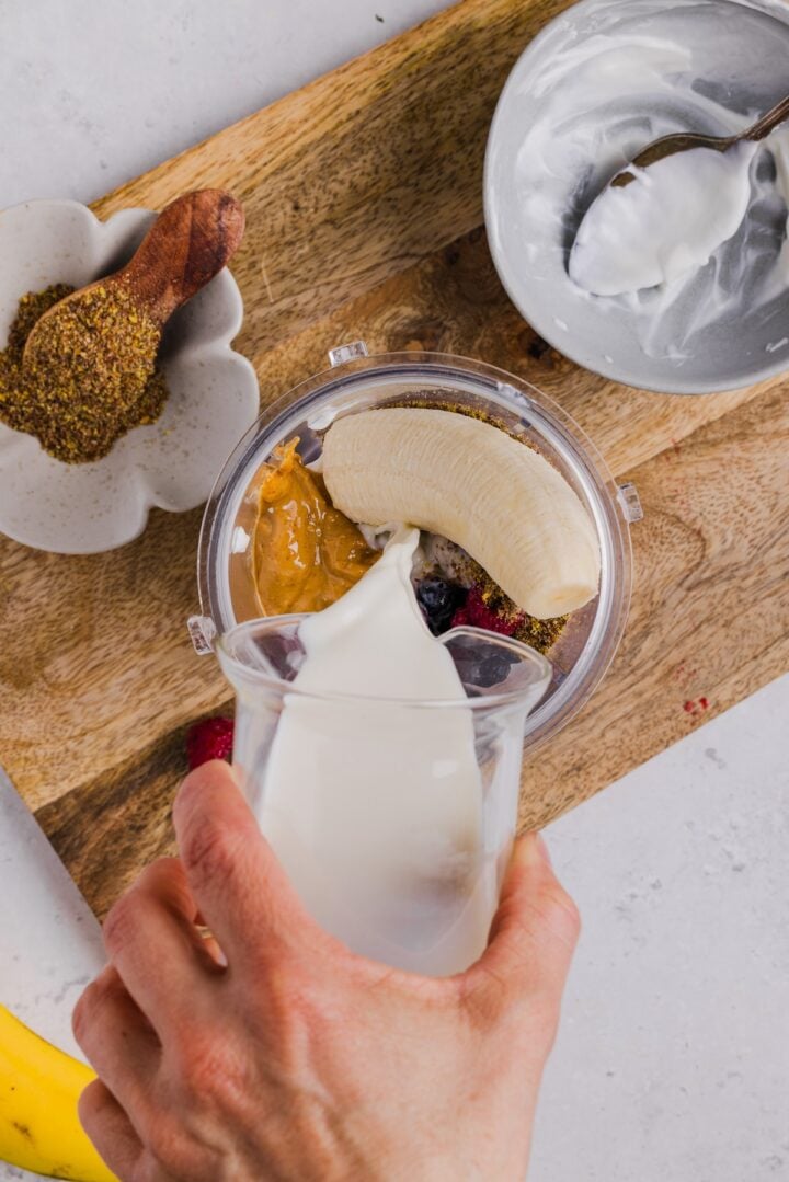 Ingredients for a mixed berry peanut butter smoothie in a blender with milk being poured in