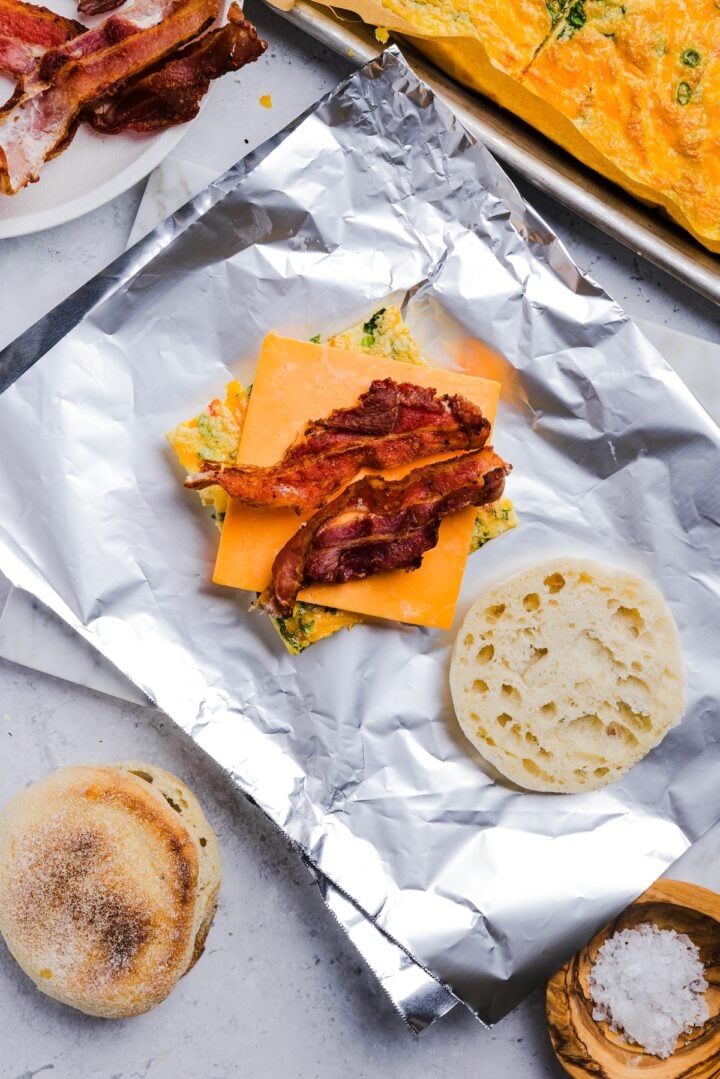 English muffin open with sheet pan eggs, bacon and a slice of cheese on aluminum foil