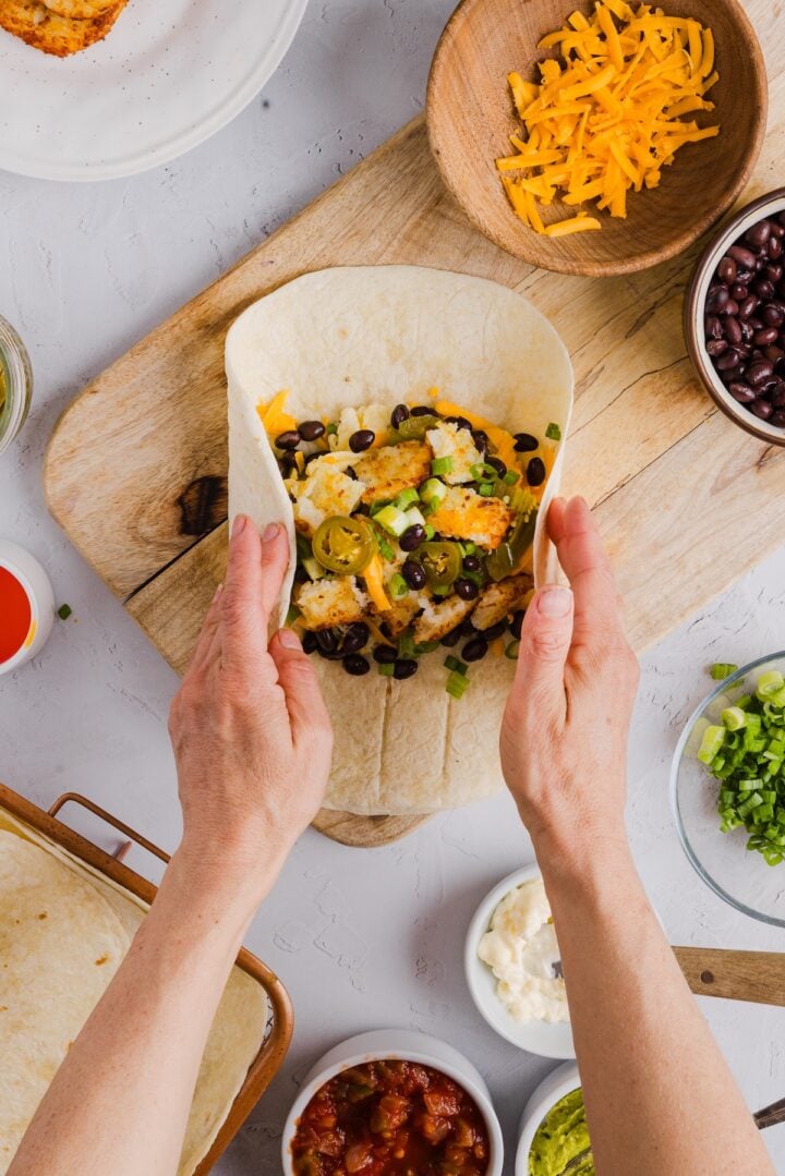 Protein breakfast burrito being wrapped by a woman's hands on a wood serving board