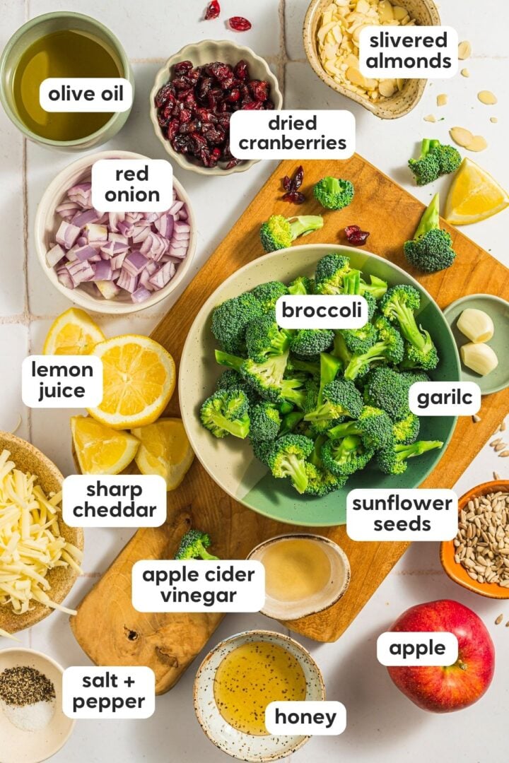 Overhead and labelled photo of ingredients in broccoli crunch salad, displayed in small bowls on a kitchen counter with a wood serving board.