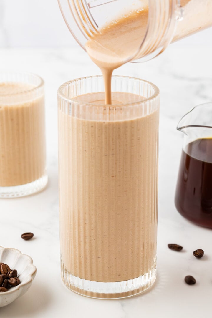 Coffee protein shake being poured into a tall etched glass on a white marble counter