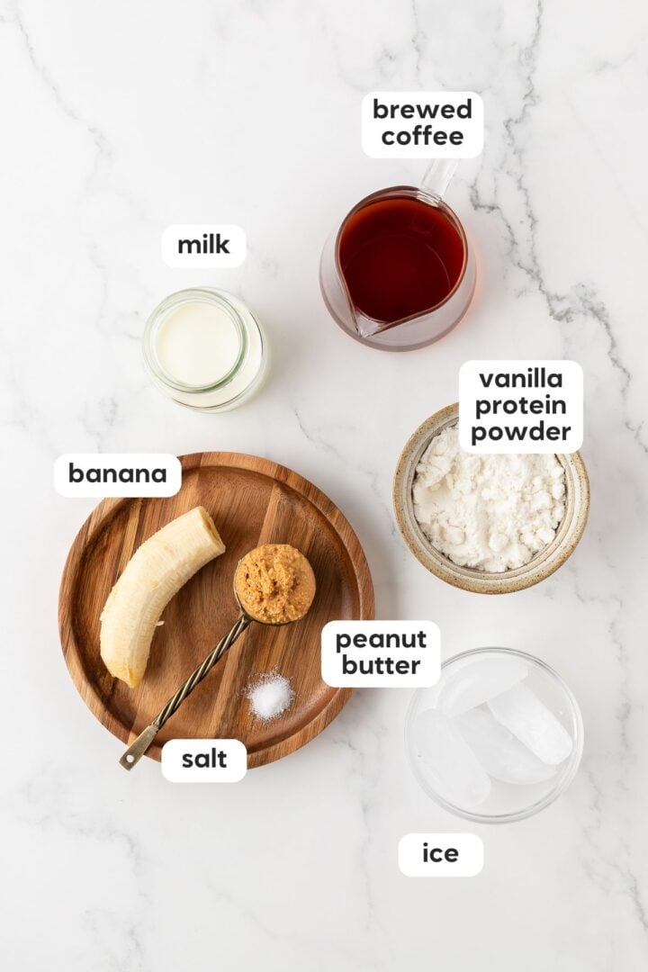 Overhead and labeled photo of ingredients in iced coffee protein shake, displayed in small bowls on a white marble counter