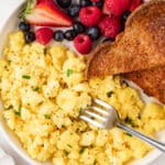 Overhead photo of cottage cheese scrambled eggs on a white plate with whole grain toast and fresh berries, garnished with chives.