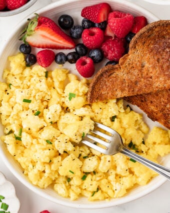 Overhead photo of cottage cheese scrambled eggs on a white plate with whole grain toast and fresh berries, garnished with chives.
