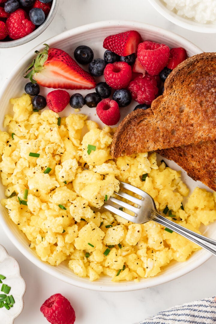 Overhead photo of cottage cheese scrambled eggs on a white plate with whole grain toast and fresh berries, garnished with chives.