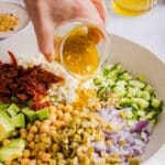 Chickpea Cucumber Feta Salad in a large, white serving bowl, with a woman's hand pouring dressing on top