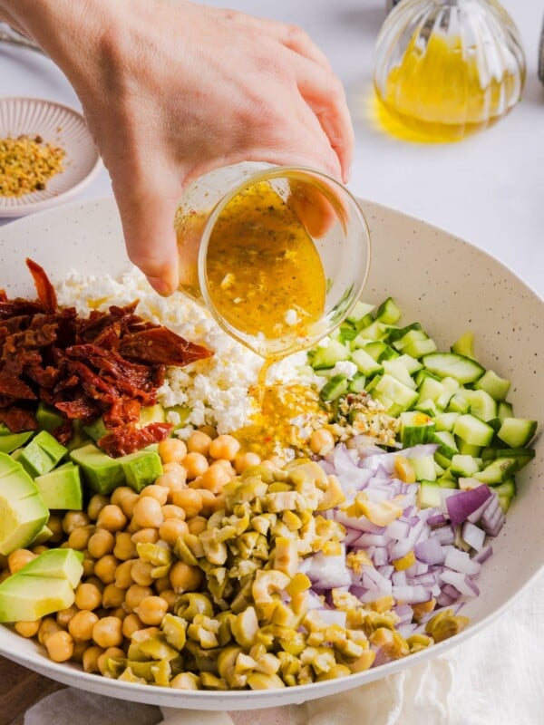 Chickpea Cucumber Feta Salad in a large, white serving bowl, with a woman's hand pouring dressing on top