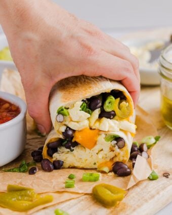 Woman's hand grabbing two halves of a high protein breakfast burrito, on a wood cutting board