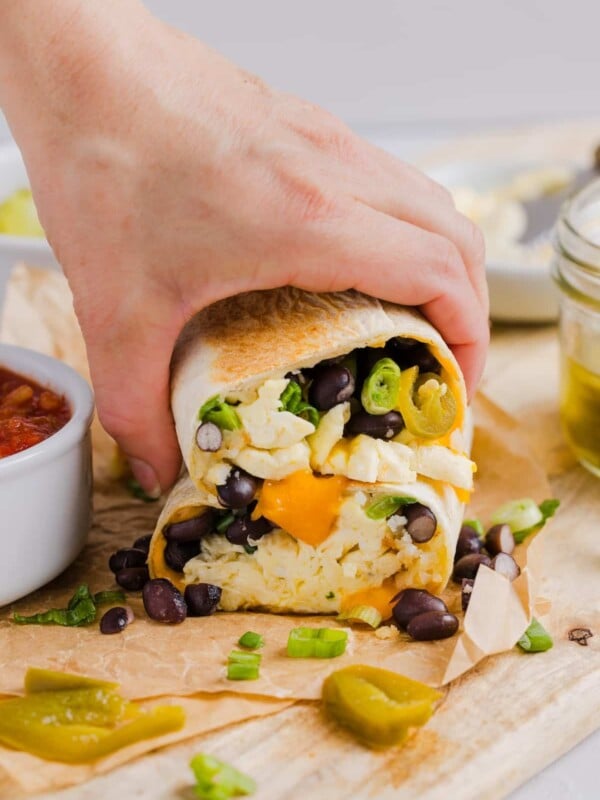 Woman's hand grabbing two halves of a high protein breakfast burrito, on a wood cutting board