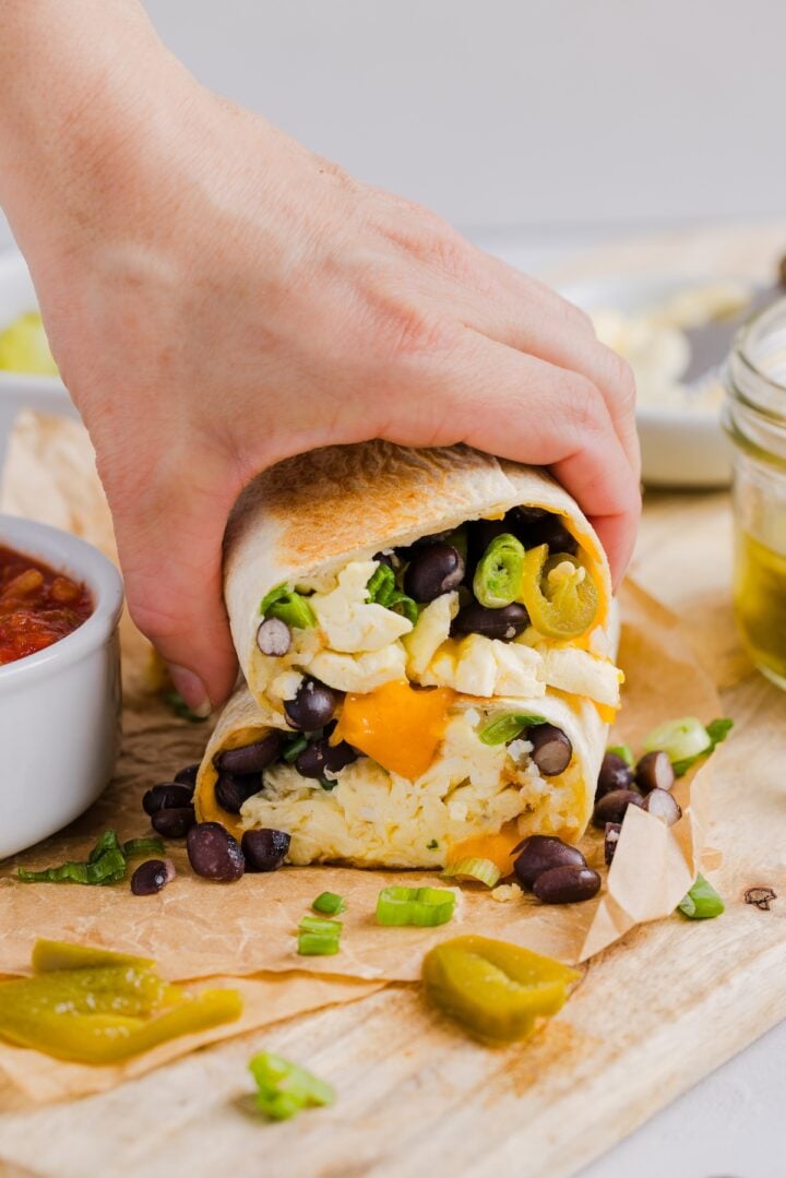 Woman's hand grabbing two halves of a high protein breakfast burrito, on a wood cutting board