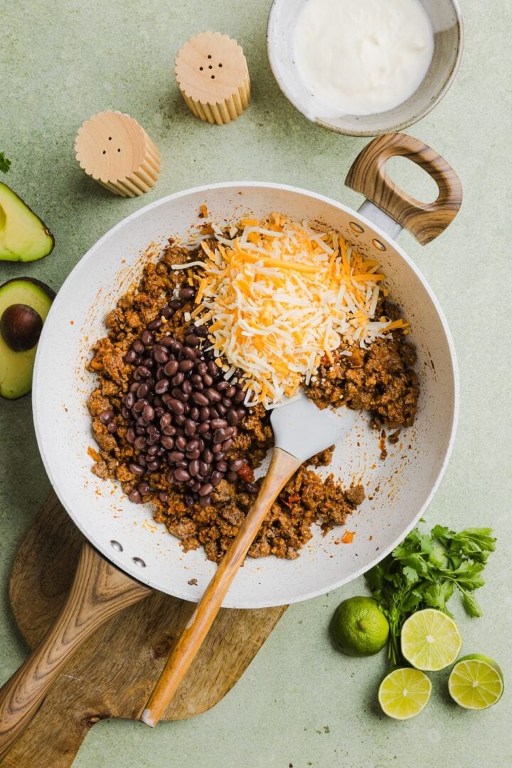 Black beans and cheese being stirred into cooked taco meat in a white pan.
