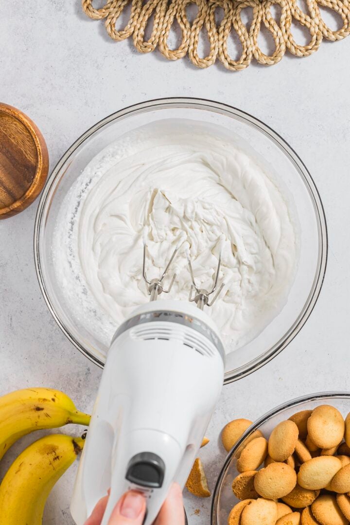 Whipping cream being blended with a hand mixer in a large glass bowl.