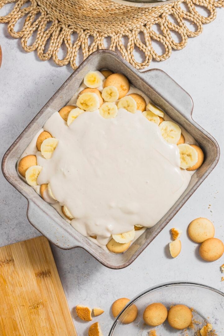 Banana pudding being assembled in a square baking dish with cookies and banana slices.