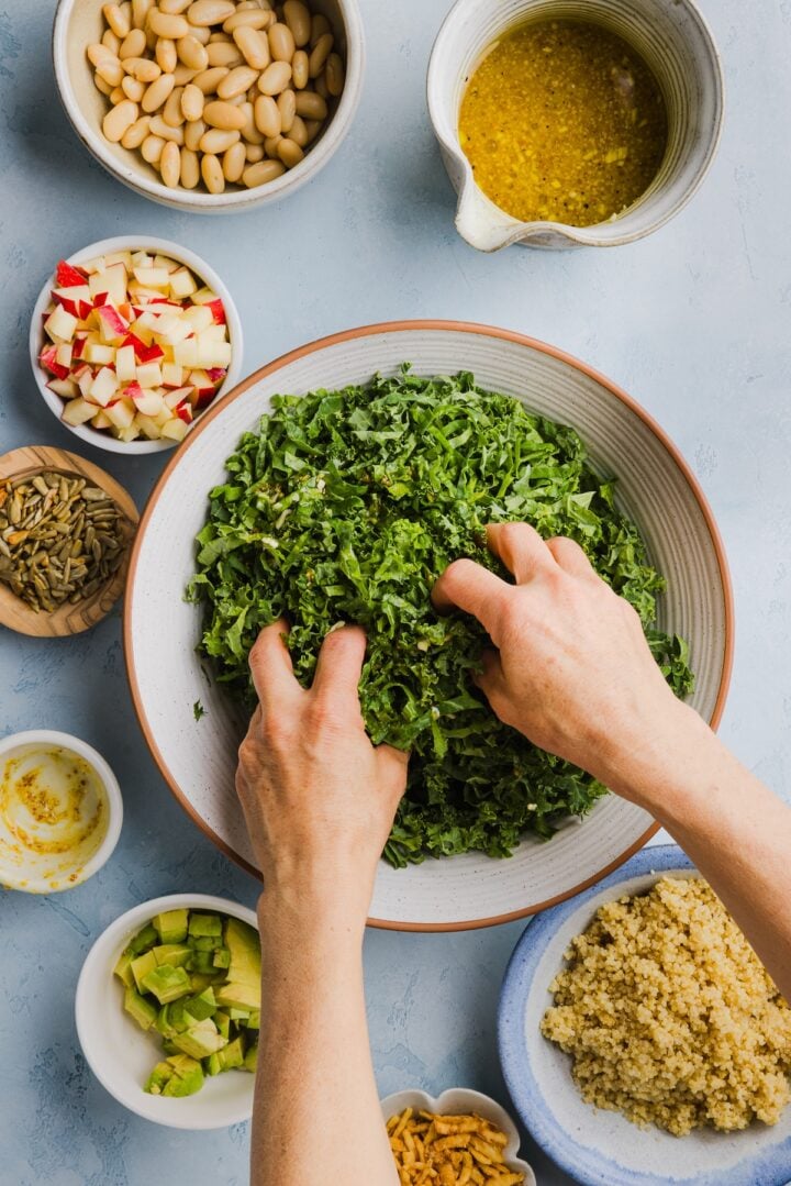 Kale being massaged with dressing in a white mixing bowl by a woman's hands.