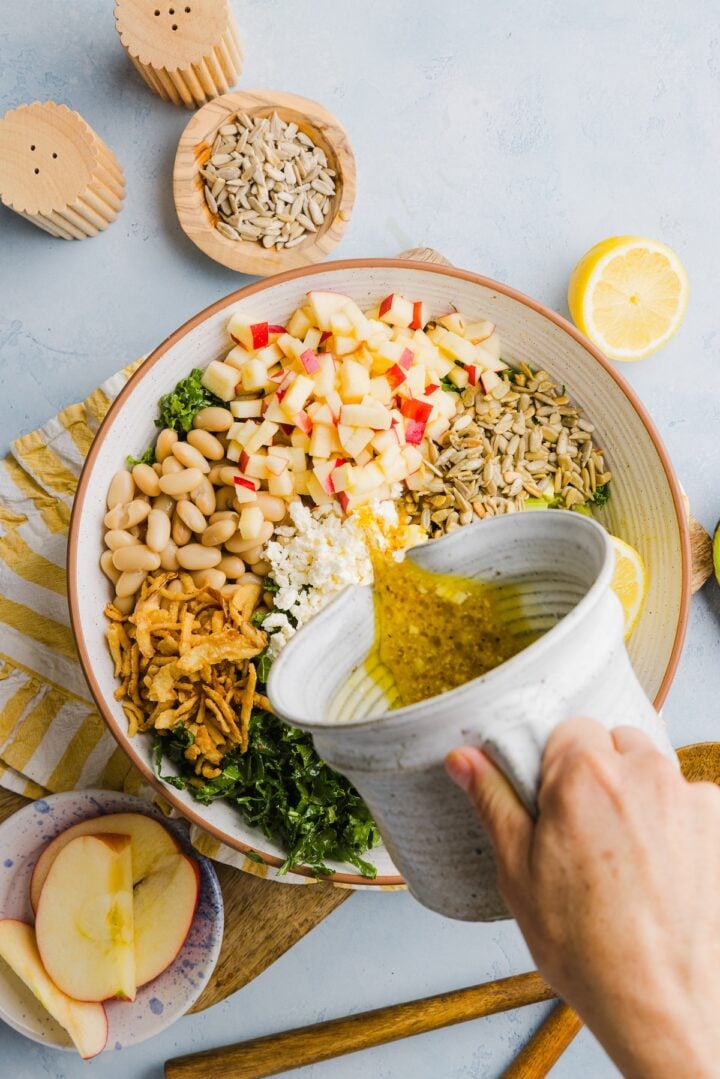 Kale white bean salad in a large white mixing bowl, with a woman's hands pouring dressing over top.