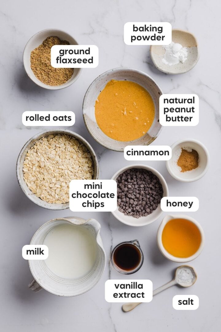 Ingredients for baked oats with chocolate chips displayed in small bowls on a white marble counter top.
