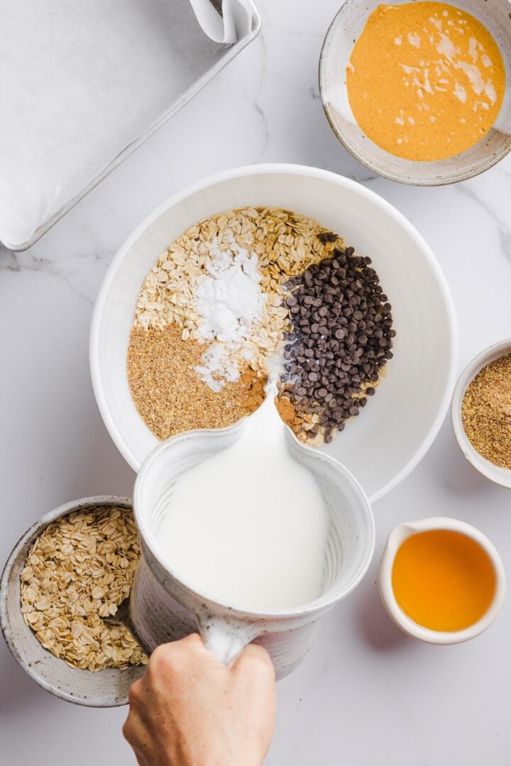 Woman's hand pouring milk into white mixing bowl with other ingredients for peanut butter baked oats.