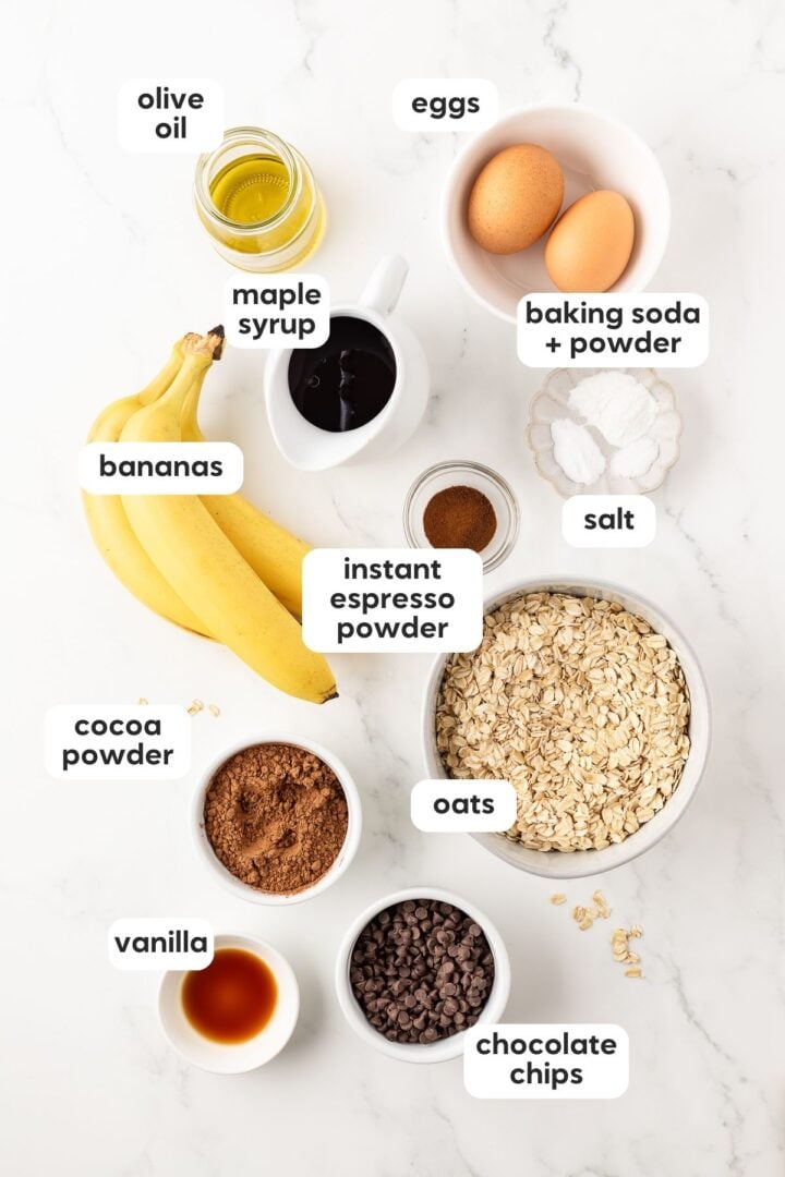 Labelled photo of ingredients for chocolate banana muffins, displayed in small bowls on a white marble counter.