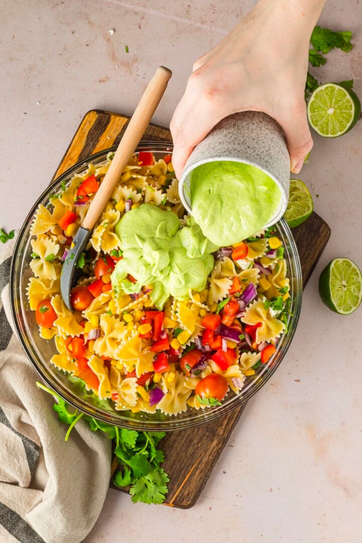 Green dressing being poured over pasta salad in a glass bowl.