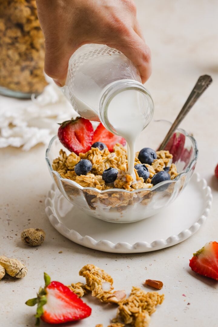 Peanut butter granola in a glass bowl with berries, with milk being poured into the bowl.