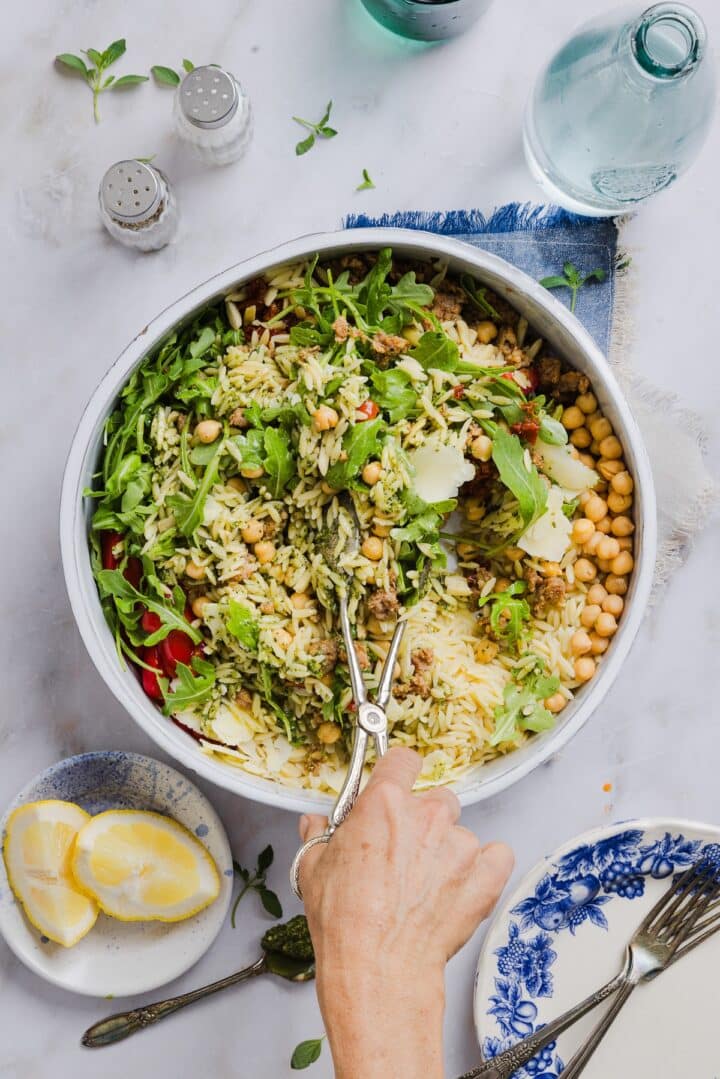 Pesto and orzo salad being tossed in a large mixing bowl by a woman's hand holding tongs.