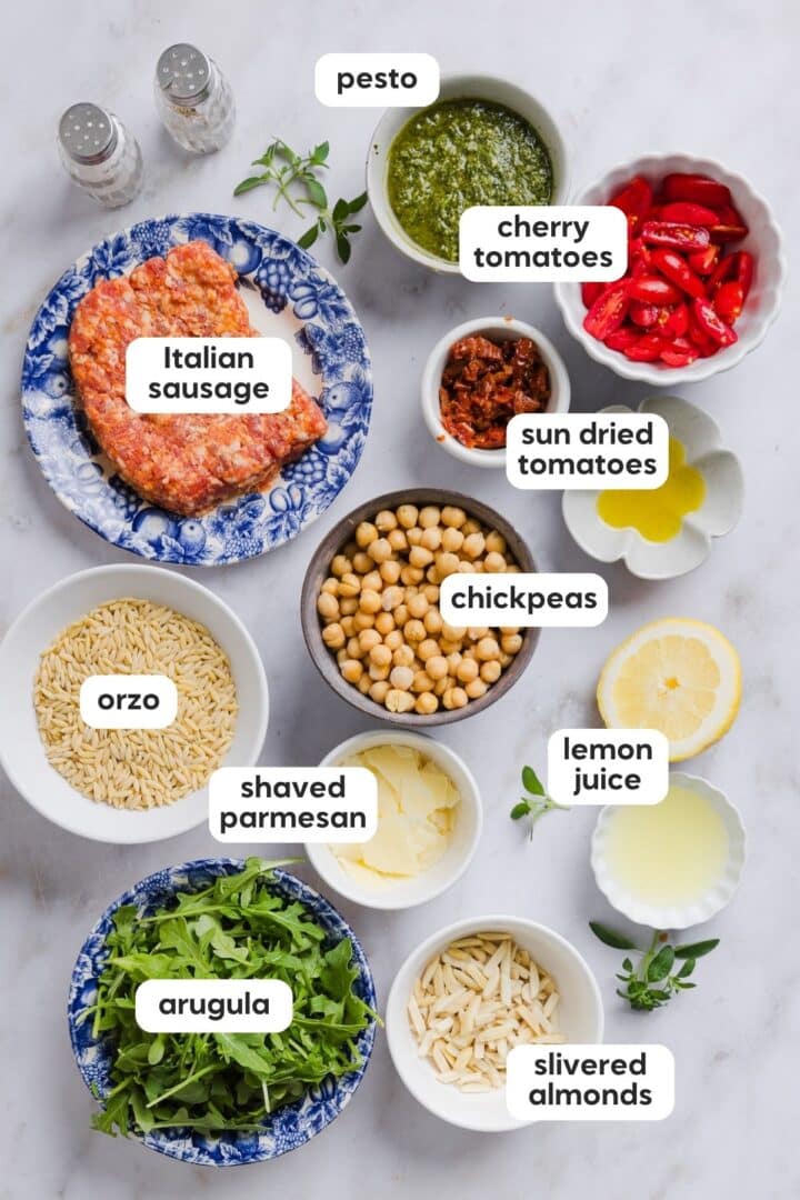 Ingredients for pesto and orzo salad with Italian sausage displayed in small bowls on a white marble counter.