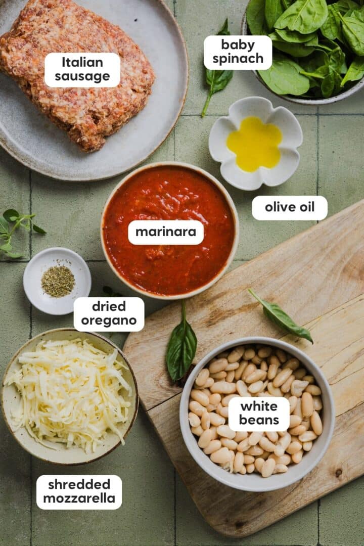 Ingredients for one-pan pizza beans and sausage displayed in small bowls on a green tile counter.