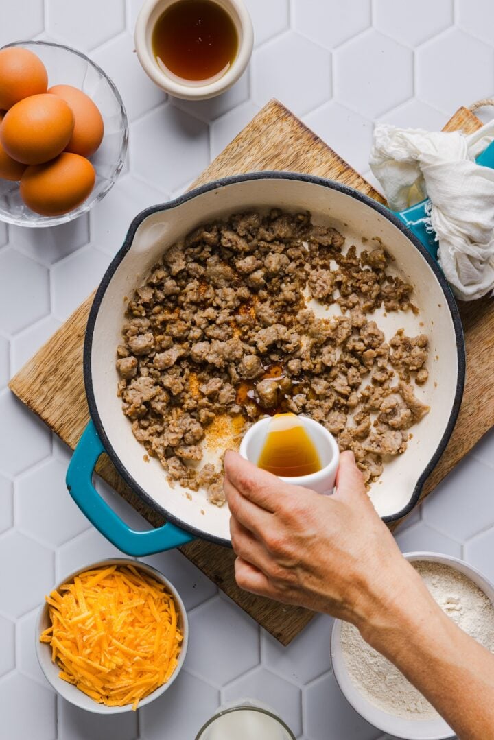 Browned and crumbled sausage in a pan with a woman's hand pouring in maple syrup.