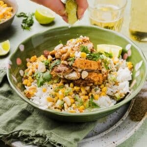 Mexican-inspired street corn chicken and rice bowl with a woman's hand squeezing a lemon wedge over top.