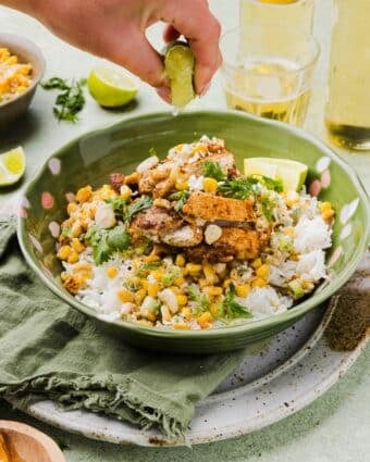 Mexican-inspired street corn chicken and rice bowl with a woman's hand squeezing a lemon wedge over top.