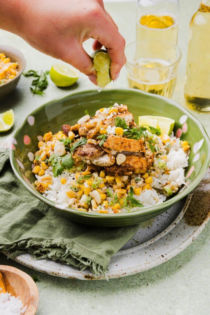 Mexican-inspired street corn chicken and rice bowl with a woman's hand squeezing a lemon wedge over top.