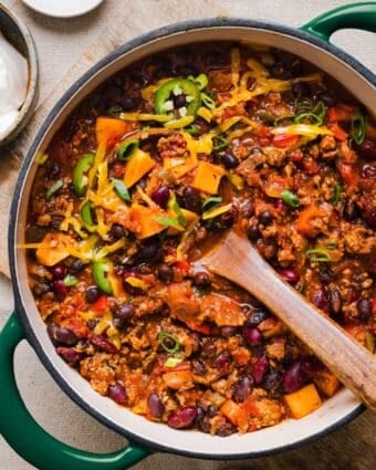 Healthy sweet potato turkey chili in a large pot, topped with shredded cheddar and green onions.