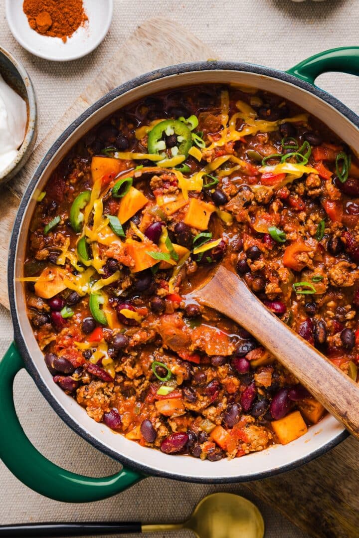 Healthy sweet potato turkey chili in a large pot, topped with shredded cheddar and green onions.