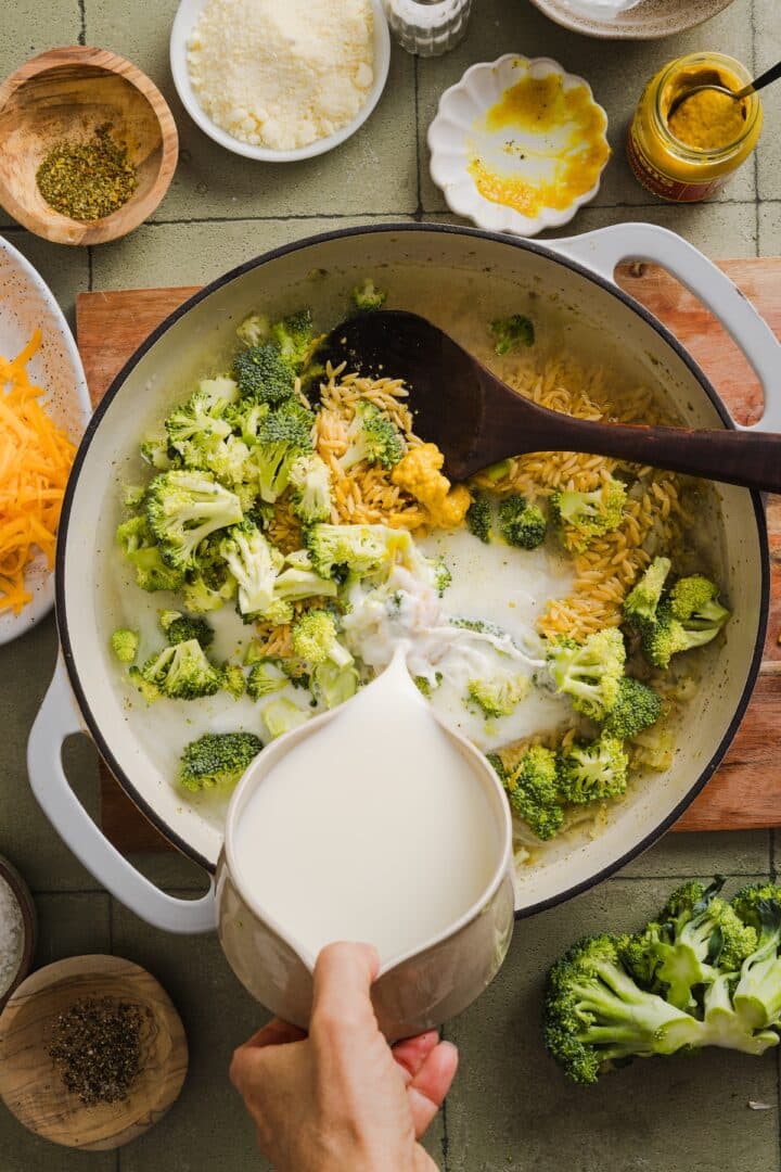 Broccoli, orzo, and milk being added to a braiser pot to make broccoli cheddar orzo.