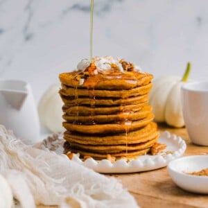 Pumpkin protein pancakes stacked on a white plate with maple syrup being poured on top.