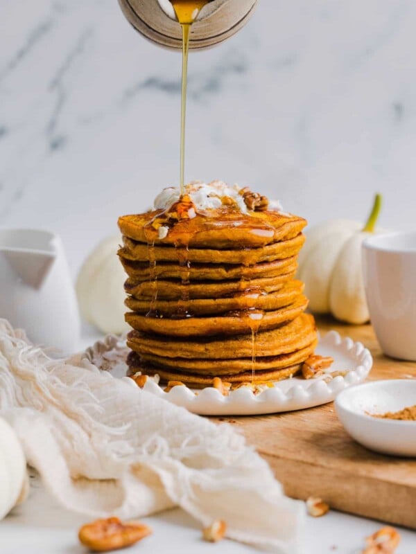 Pumpkin protein pancakes stacked on a white plate with maple syrup being poured on top.