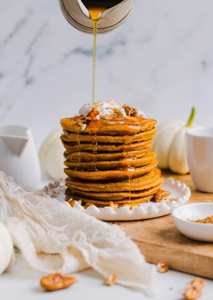 Pumpkin protein pancakes stacked on a white plate with maple syrup being poured on top.