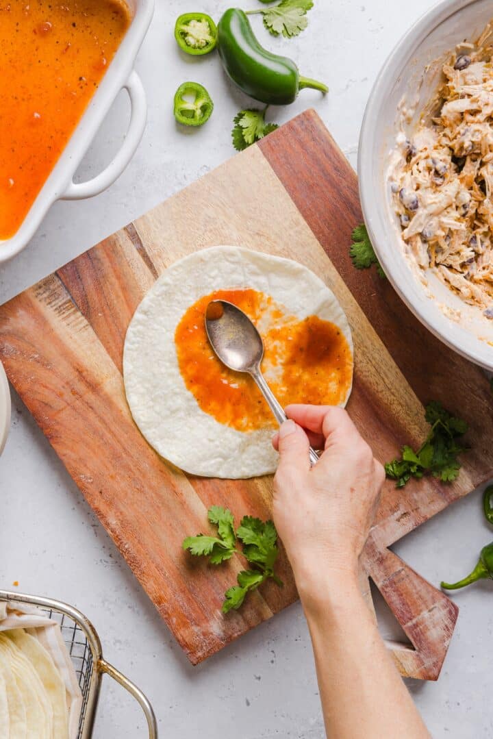 Enchilada sauce being spooned over a small tortilla on a wood cutting board.