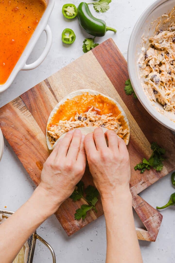 Chicken enchilada being rolled on a wooden cutting board by a woman's hand.