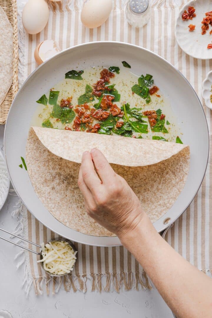Woman's hand placing a whole wheat tortilla over egg whites in a large ceramic pan.