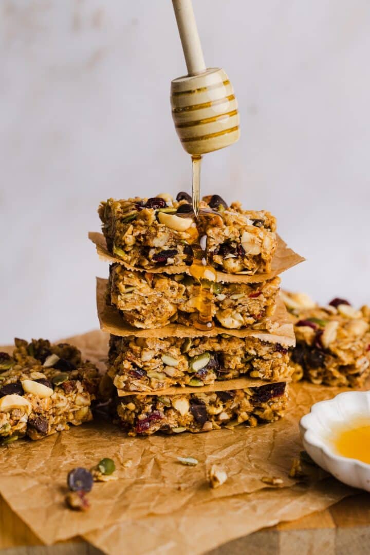 Three no-bake trail mix granola bars stacked on parchment paper, with a drizzle of honey coming down over top.