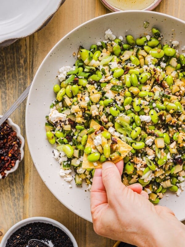 Cucumber edamame salad in a large white bowl, with a woman's hand scooping some with a tortilla chip.