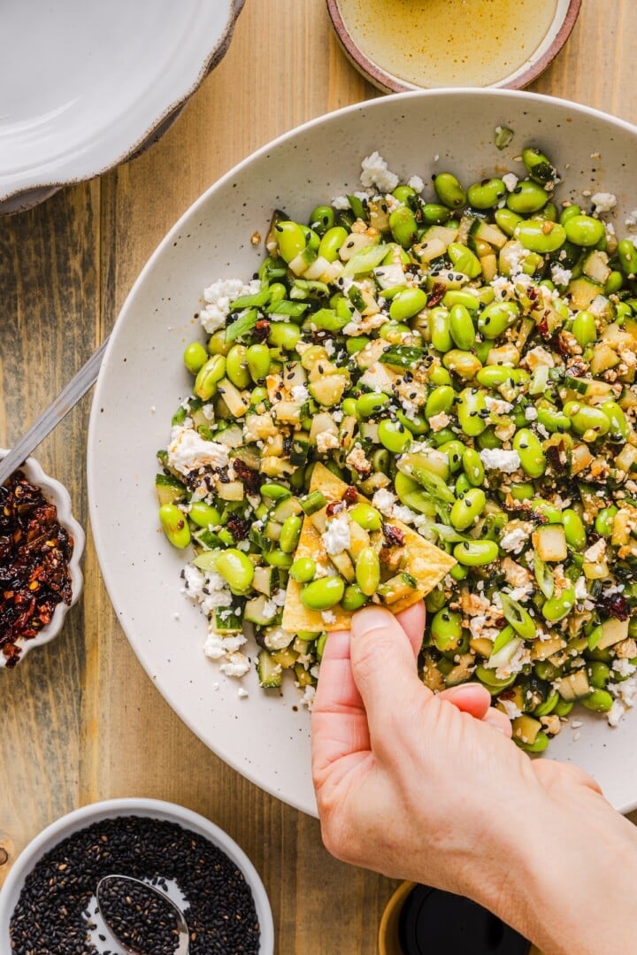 Cucumber edamame salad in a large white bowl, with a woman's hand scooping some with a tortilla chip.