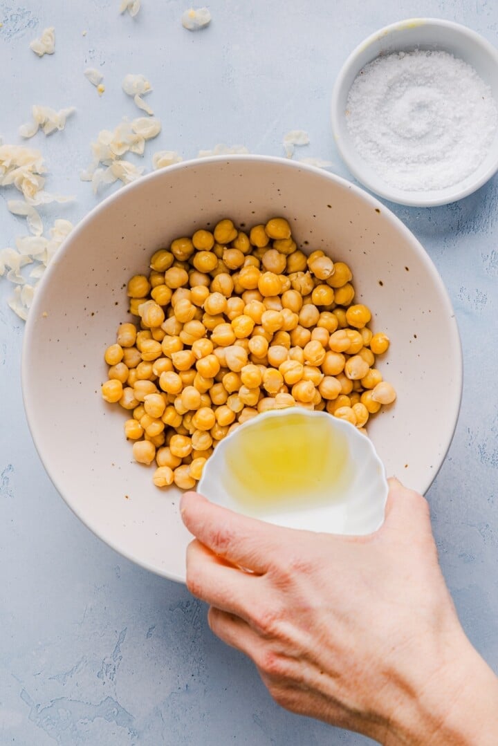 Woman's hand pouring oil over canned chickpeas in a large, white mixing bowl.