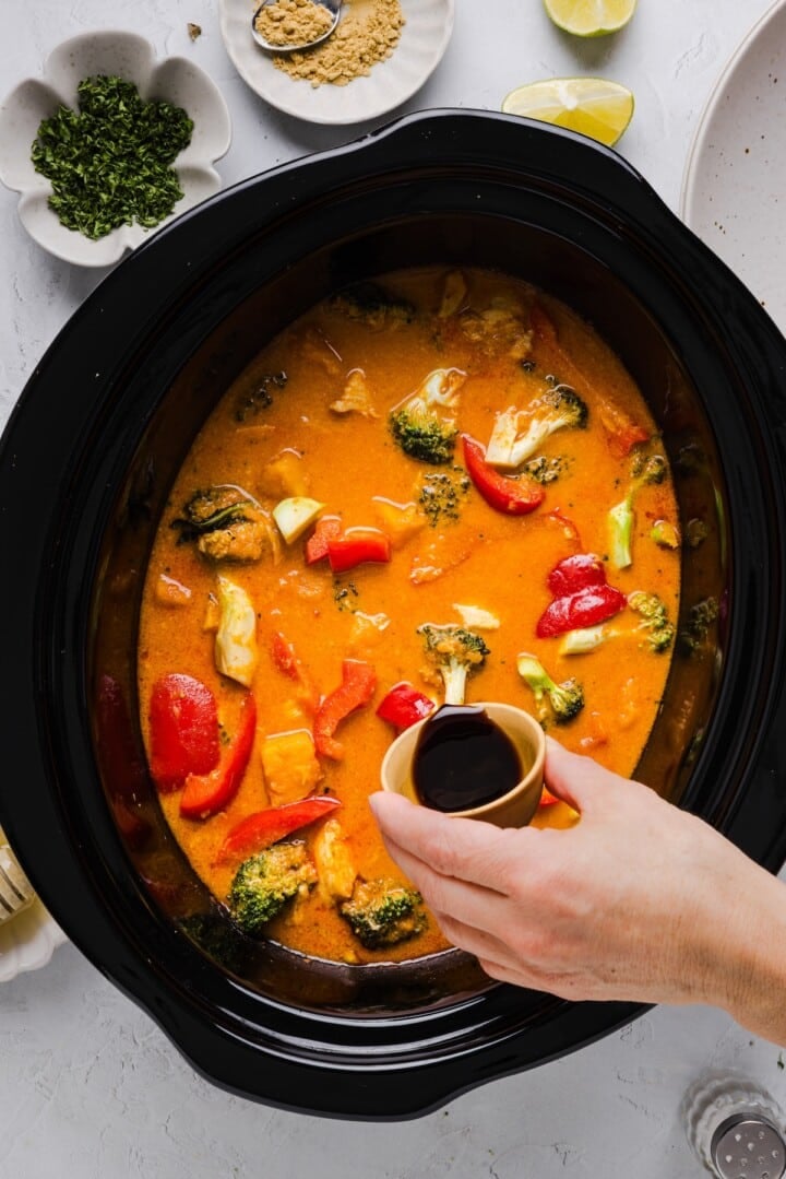Slow cooker thai-inspired red curry in a slow cooker, with a woman's hand pouring in soy sauce.