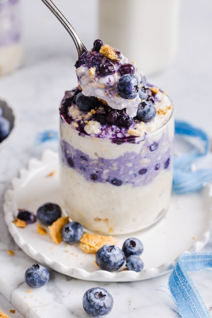 Blueberry cheesecake overnight oats in a glass jar, with a spoonful held over top.